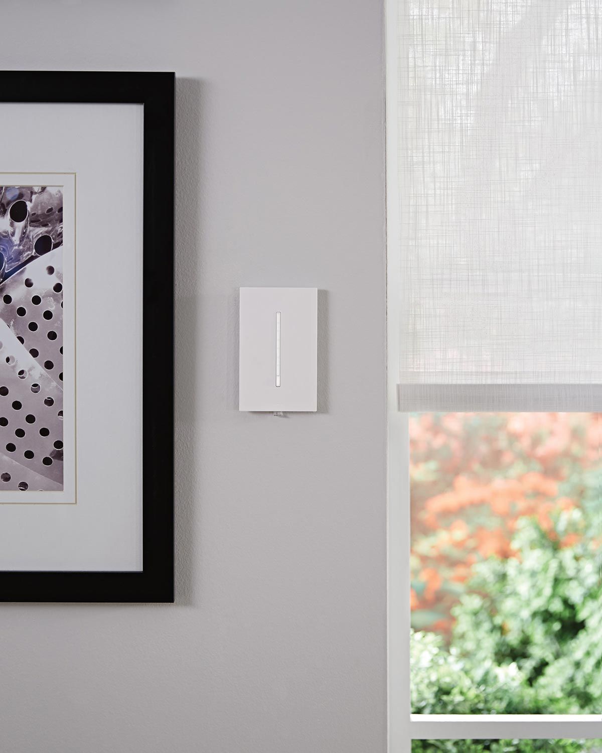 A Lutron control panel on a gray wall next to a modern art piece and a window with sheer white blinds, highlighting the sleek integration of smart home technology.